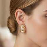 A close-up side profile of a woman wearing a gold-toned triple-row pearl stud earring, with her hair in a low bun against a softly blurred indoor background.