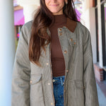 A woman wearing a brown and beige houndstooth jacket with brown collar and button details, a brown mock neck top, and blue jeans, standing in front of a storefront.