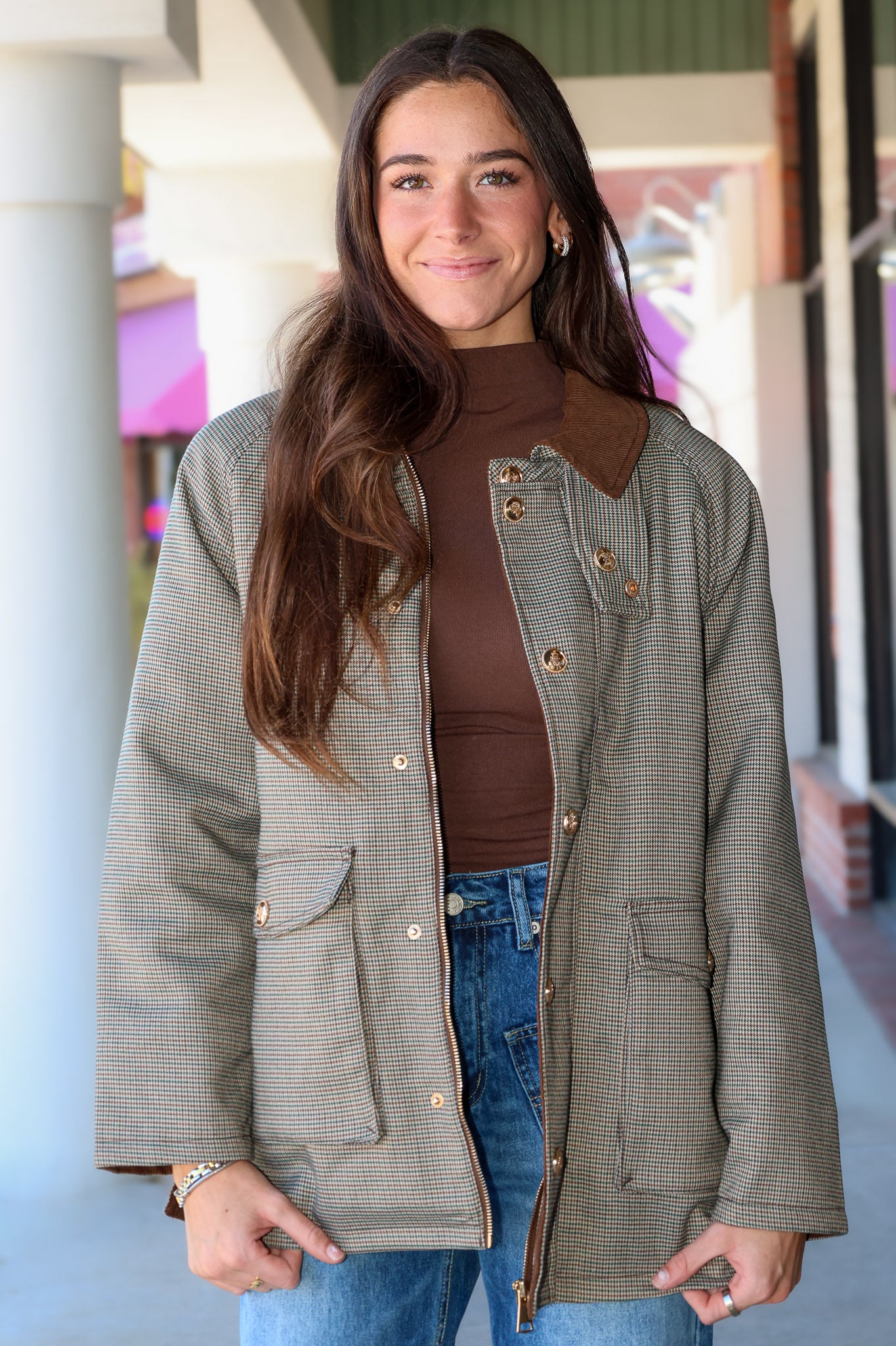 A woman wearing a brown and beige houndstooth jacket with brown collar and button details, a brown mock neck top, and blue jeans, standing in front of a storefront.