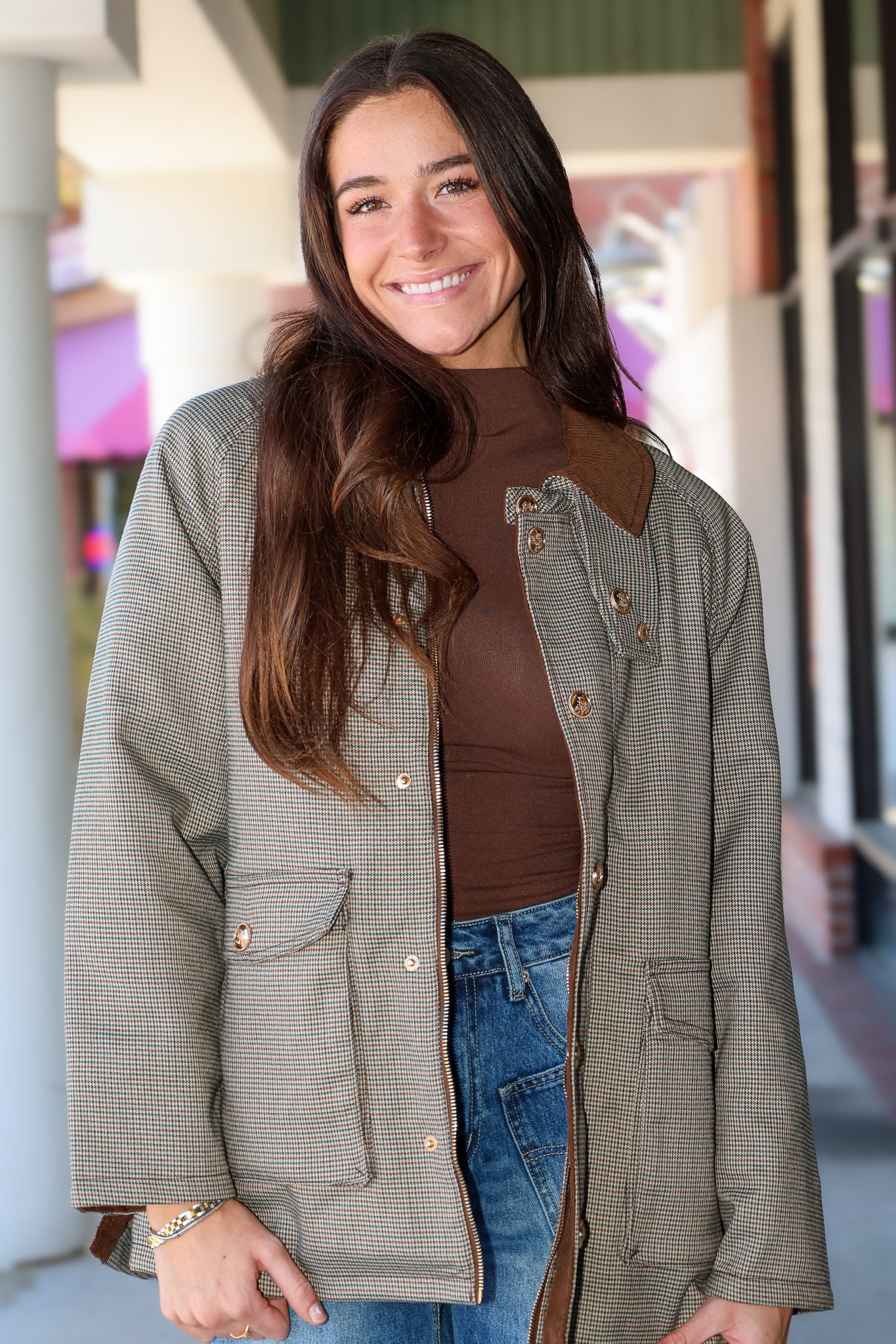 A woman smiling while wearing a brown and beige houndstooth patterned utility jacket with brown leather-like collar and pocket trim, layered over a brown top and blue jeans.