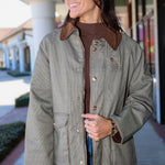 A woman wearing a brown and gray micro-check patterned jacket with a brown corduroy collar, styled over a brown turtleneck and blue jeans, standing outdoors in front of a storefront.