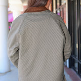 A person viewed from behind wearing a textured, small-scale checkered olive green jacket with a brown collar, standing in an outdoor walkway.