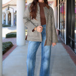 A woman stands on a covered walkway wearing a light olive-gray quilted jacket over a brown mock-neck top, blue wide-leg jeans, and white heeled sandals.