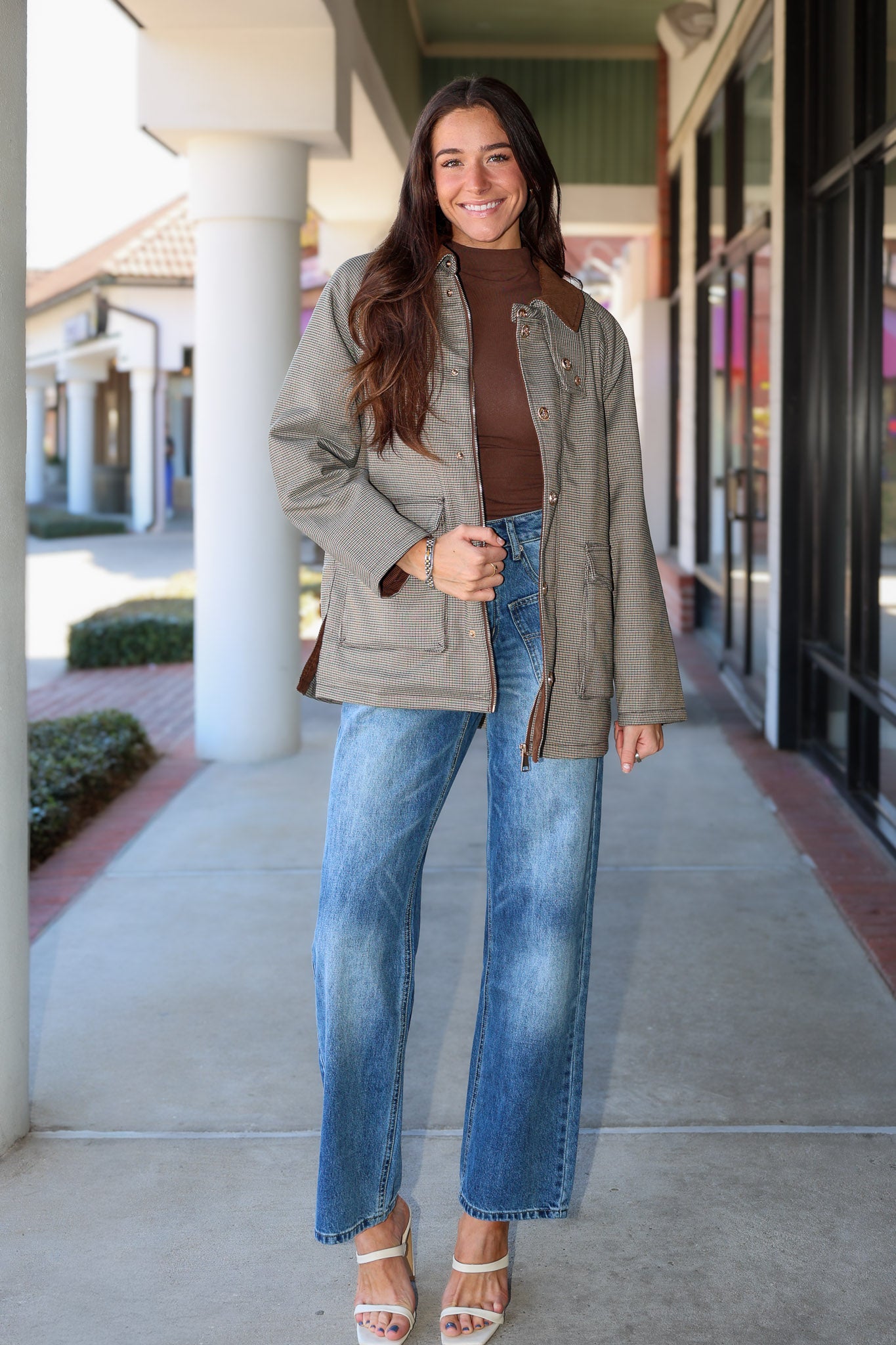 A woman stands on a covered walkway wearing a light olive-gray quilted jacket over a brown mock-neck top, blue wide-leg jeans, and white heeled sandals.