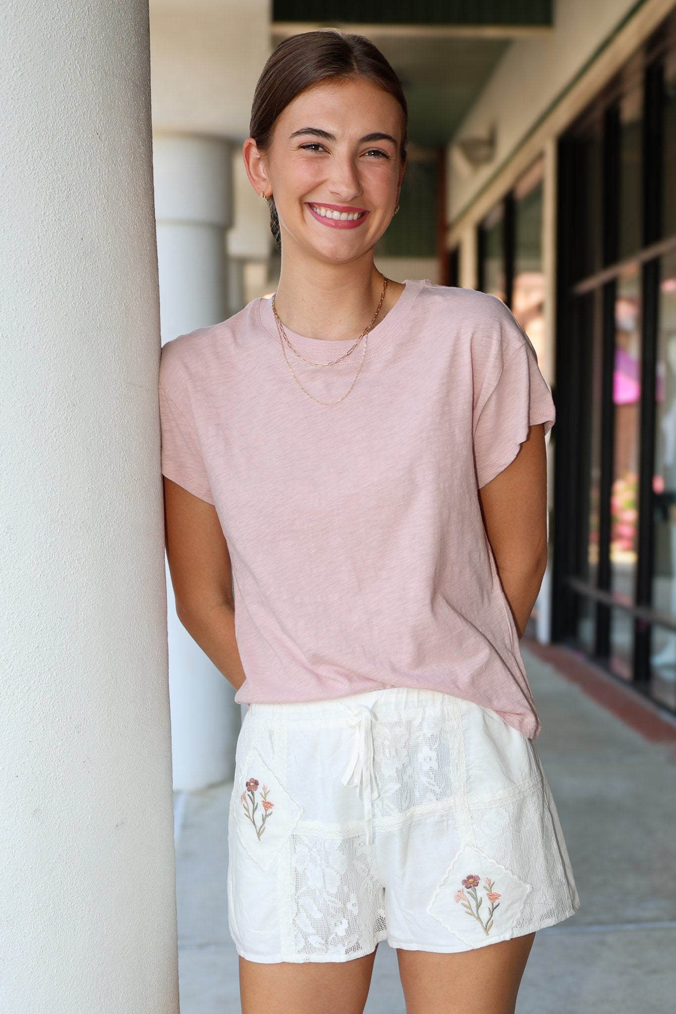 Woman wearing a pink top and white shorts with floral embroidery, standing against a building exterior.