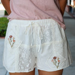 Person wearing a pink top and white lace shorts with floral embroidery on a blurred outdoor background