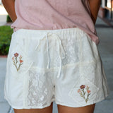 Person wearing a pink top and white lace shorts with floral embroidery on a blurred outdoor background