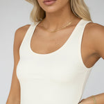 A woman wearing a ribbed cream-colored tank top and white pants, shown from the shoulders to the hips against a plain light gray background.