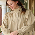 Woman wearing a khaki jacket and white pants standing outdoors.