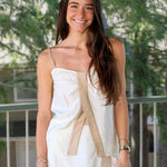 A woman with long brown hair stands outdoors wearing a white sleeveless mini dress with beige tie-front panels and beige hem trim. She is smiling and posing in front of a railing with green trees in the background.