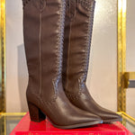 A pair of Finley knee-high brown leather boots with decorative braided stitching near the top and stacked on a bright pink book titled 'Christian Louboutin Exhibition'.