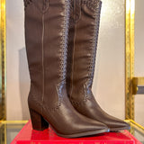 A pair of Finley knee-high brown leather boots with decorative braided stitching near the top and stacked on a bright pink book titled 'Christian Louboutin Exhibition'.
