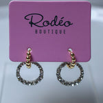 A pair of gold hoop earrings with sparkling clear stones, displayed on a pink card labeled 'Rodéo BOUTIQUE'.