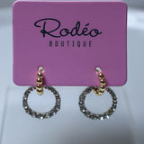 A pair of gold hoop earrings with sparkling clear stones, displayed on a pink card labeled 'Rodéo BOUTIQUE'.