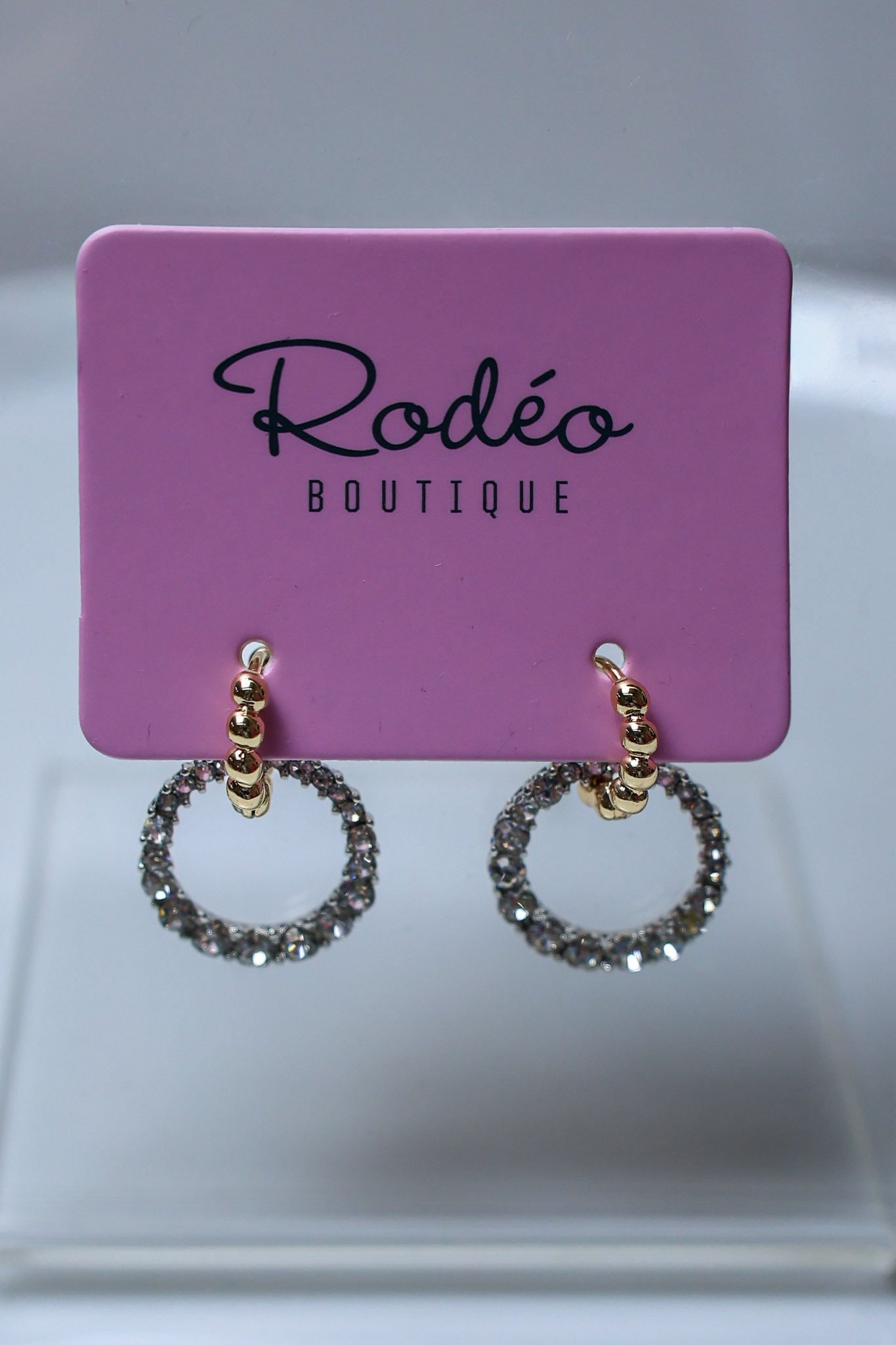 A pair of gold hoop earrings with sparkling clear stones, displayed on a pink card labeled 'Rodéo BOUTIQUE'.
