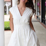 Woman wearing a white cap sleeve bubble  hem dress with pearl detail standing outdoors on a sidewalk.