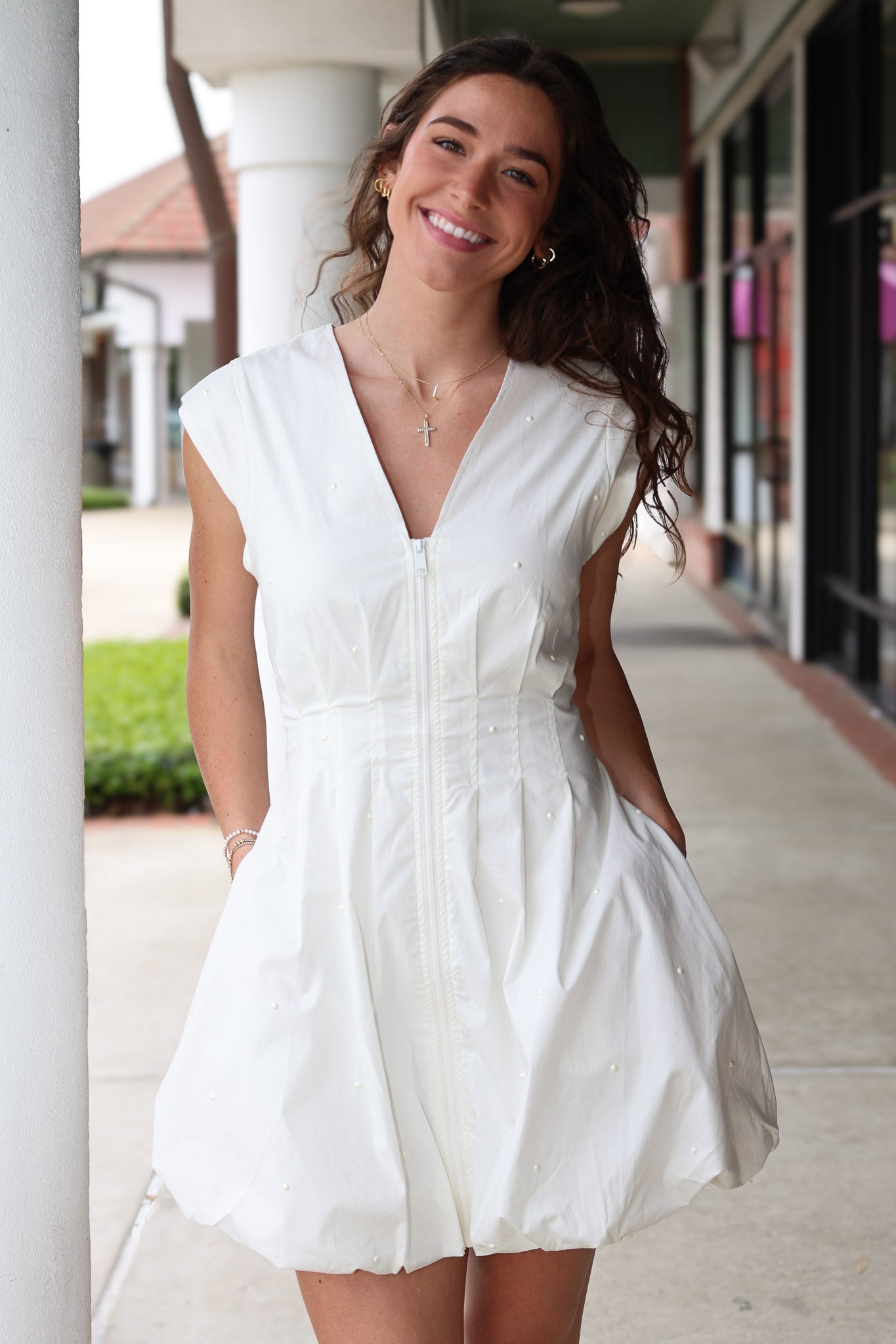 Woman wearing a white cap sleeve bubble  hem dress with pearl detail standing outdoors on a sidewalk.