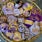 A gold-colored tray filled with various round pinback buttons featuring purple, yellow, black, and white designs, including tiger graphics and text such as 'LANE TRAIN', 'TIGER TERRITORY', and 'HEAUX'.