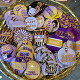 A gold-colored tray filled with various round pinback buttons featuring purple, yellow, black, and white designs, including tiger graphics and text such as 'LANE TRAIN', 'TIGER TERRITORY', and 'HEAUX'.