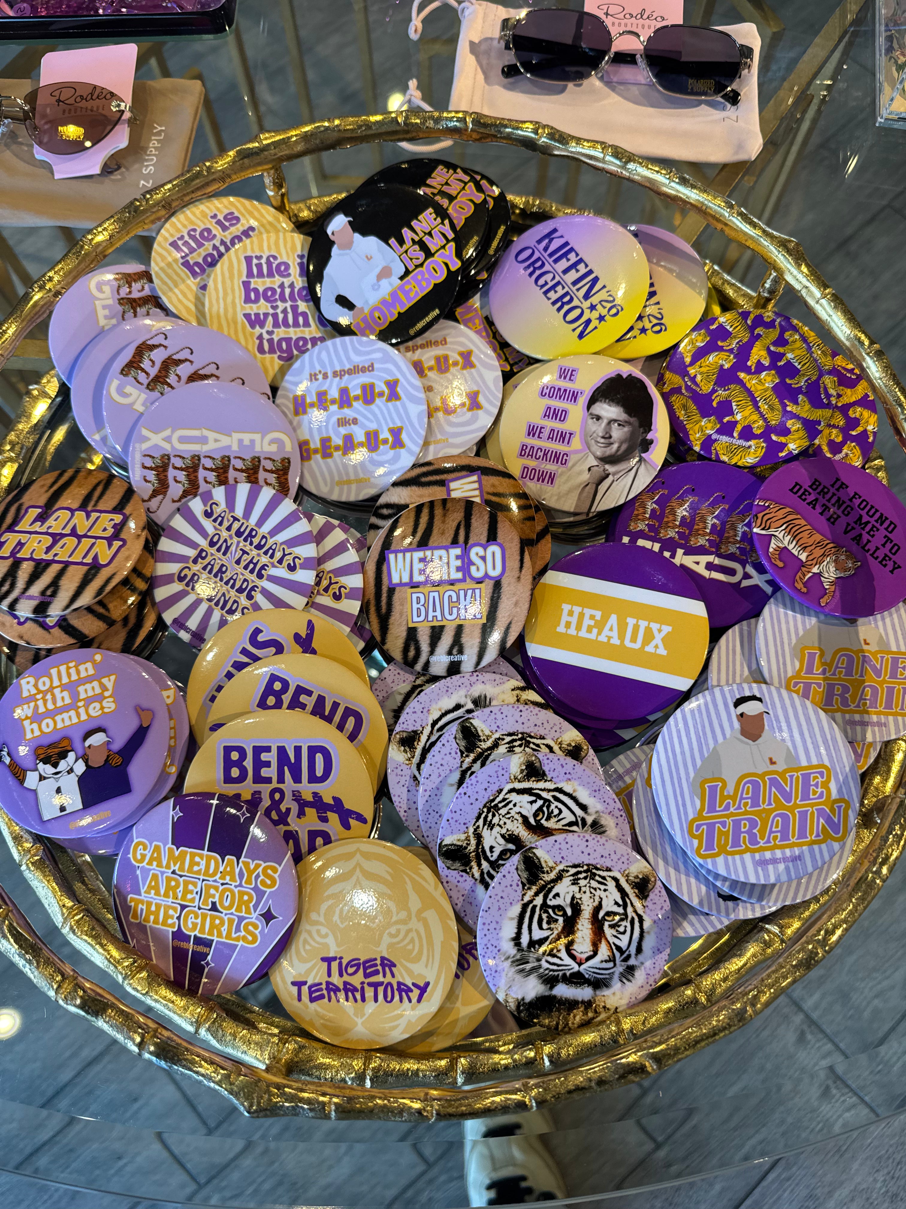 A gold-colored tray filled with various round pinback buttons featuring purple, yellow, black, and white designs, including tiger graphics and text such as 'LANE TRAIN', 'TIGER TERRITORY', and 'HEAUX'.