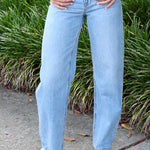 Woman wearing a denim vest and jeans standing outdoors with greenery and a yellow trailer in the background.