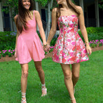 Two women in pink dresses standing on grass with a garden and building in the background
