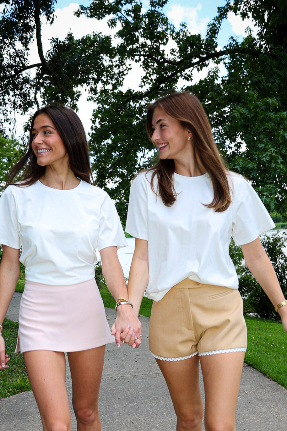 Two women walking hand-in-hand on a path with trees and a lake in the background.