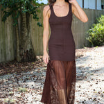A woman wearing a brown sleeveless knit dress with a sheer mesh overlay skirt, standing outdoors on a gravel path with a wooden fence and trees in the background.
