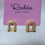 A pair of gold-toned arch-shaped stud earrings covered in small clear rhinestones, displayed on a pink Rodéo Boutique card against a light gray background.