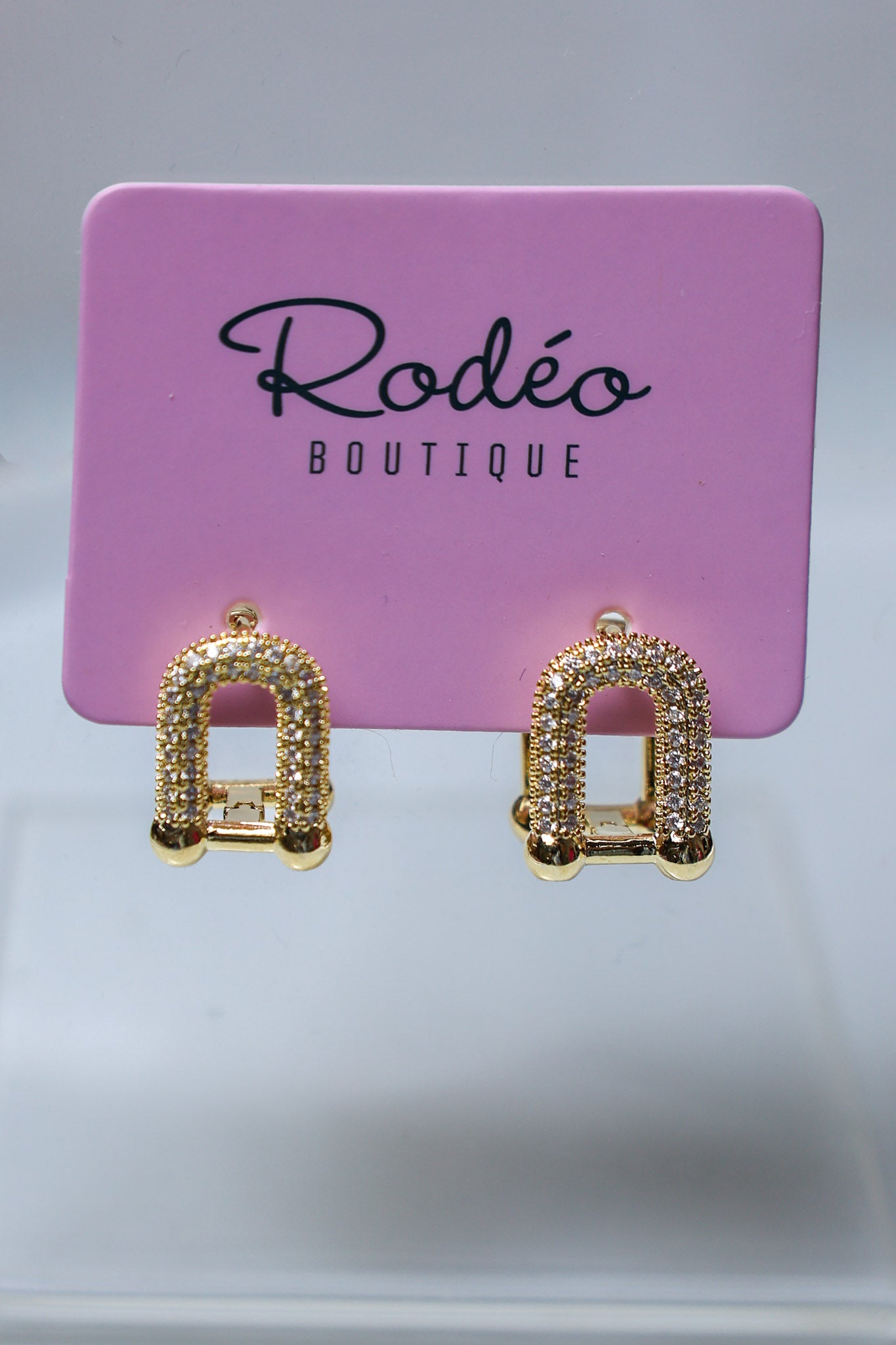 A pair of gold-toned arch-shaped stud earrings covered in small clear rhinestones, displayed on a pink Rodéo Boutique card against a light gray background.