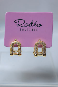 A pair of gold-toned arch-shaped stud earrings covered in small clear rhinestones, displayed on a pink Rodéo Boutique card against a light gray background.