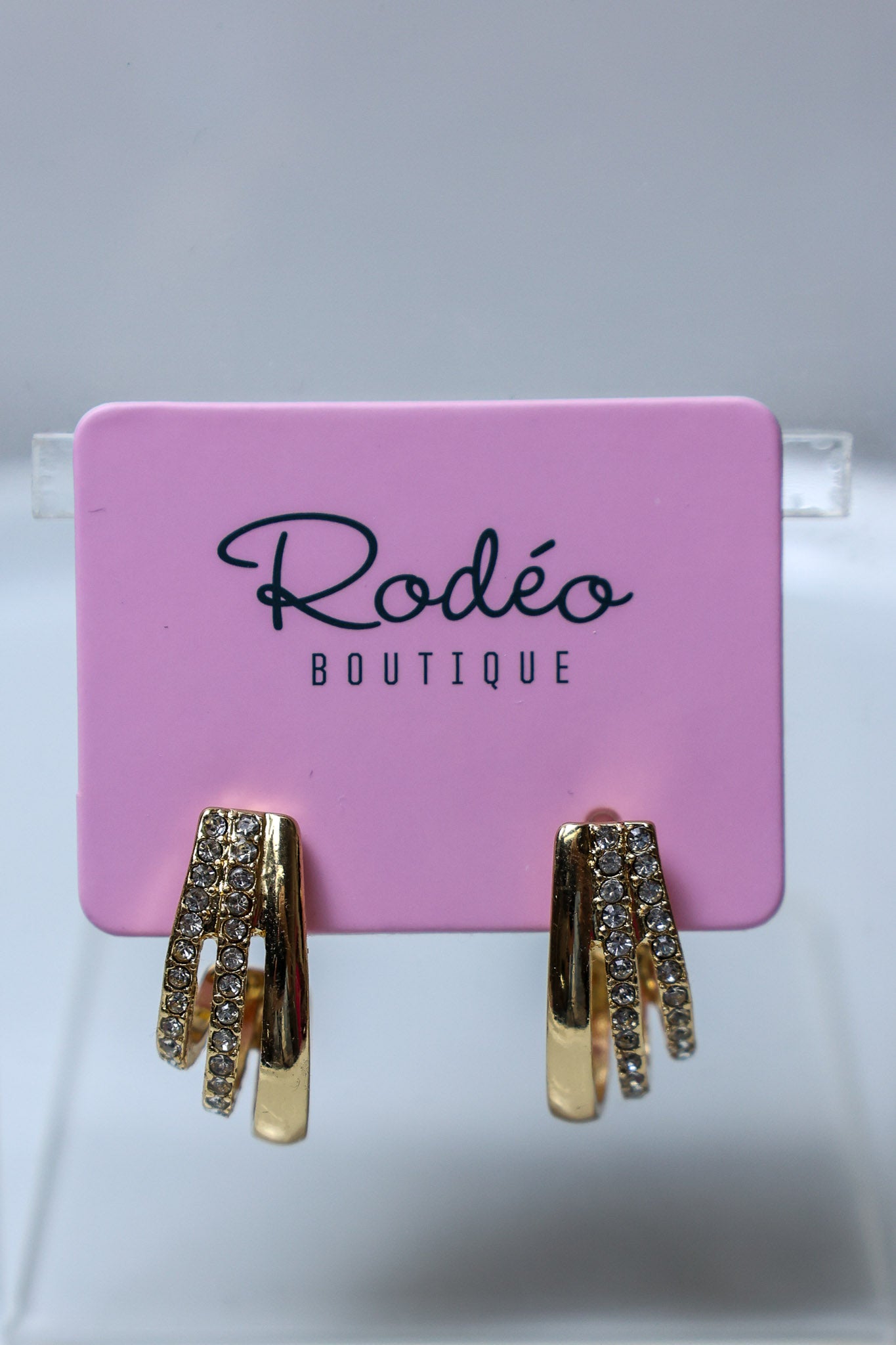 Gold huggie hoop earrings with clear rhinestone accents, displayed on a pink Rodéo Boutique card.