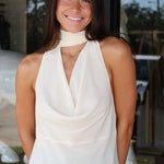 A smiling woman with long dark hair wearing a cream-colored halter top with a high neck and draped front, paired with blue jeans, standing in front of a glass storefront.