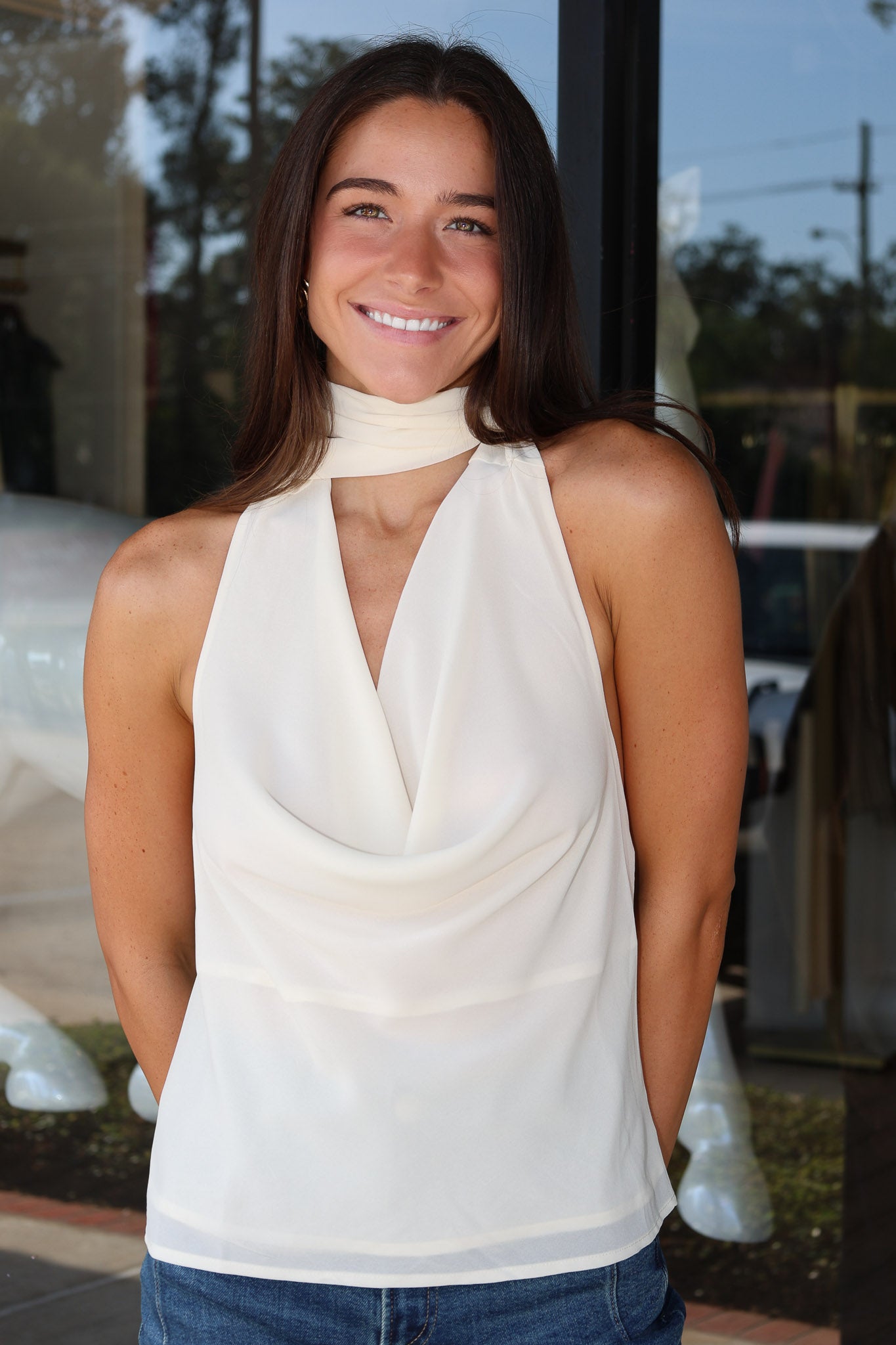 A smiling woman with long dark hair wearing a cream-colored halter top with a high neck and draped front, paired with blue jeans, standing in front of a glass storefront.
