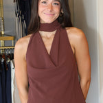 A woman wearing a sleeveless, high-neck halter top in a dusty rose-brown color, paired with blue jeans, standing in front of a clothing rack.