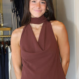 A woman wearing a sleeveless, high-neck halter top in a dusty rose-brown color, paired with blue jeans, standing in front of a clothing rack.