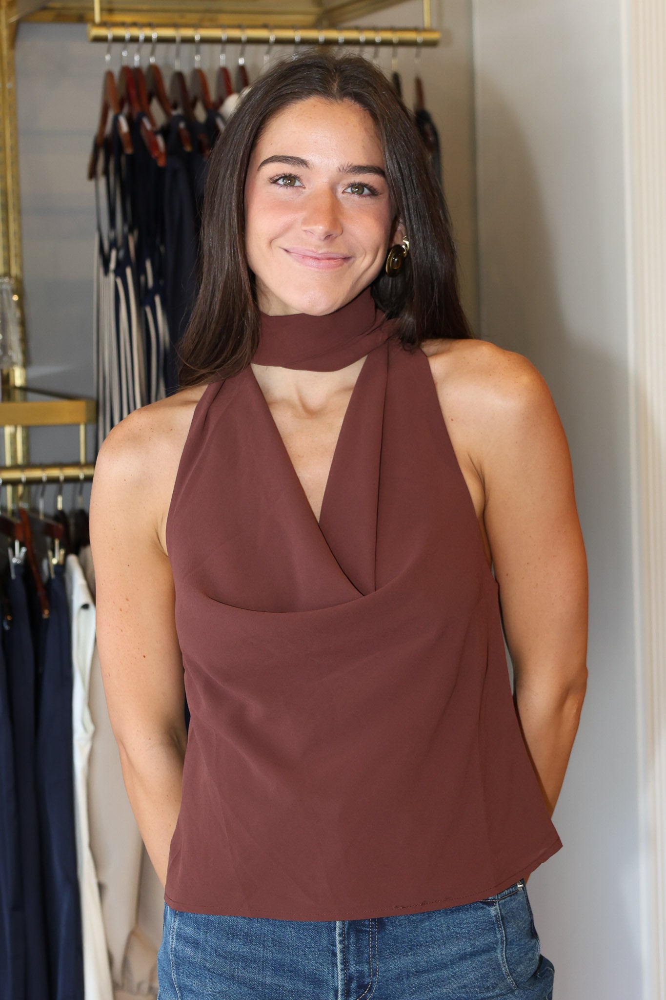A woman wearing a sleeveless, high-neck halter top in a dusty rose-brown color, paired with blue jeans, standing in front of a clothing rack.