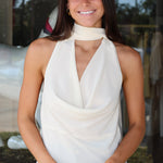 A woman smiling and wearing a sheer white halter-neck top with a draped cowl neckline, paired with blue jeans.