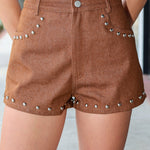 A person wearing brown denim shorts with silver stud embellishments along the pocket edges and hem, paired with a white top.