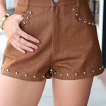 A person wearing brown studded shorts with silver metal studs along the front pockets and hem, paired with a white top. The photo is cropped from the chest down, showing the shorts being worn in a real-world setting.