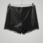 Black faux leather shorts with a floral lace hem, hanging from a wooden hanger with metal clips against a textured beige wall.