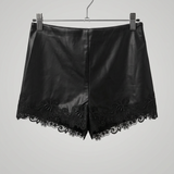 Black faux leather shorts with a floral lace hem, hanging from a wooden hanger with metal clips against a textured beige wall.