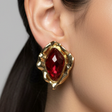 Gold earring with a red gemstone worn by a person against a neutral background