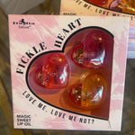 Heart-shaped lip oils in a box with 'Fickle Heart' branding on a reflective surface.
