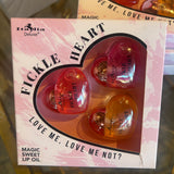 Heart-shaped lip oils in a box with 'Fickle Heart' branding on a reflective surface.
