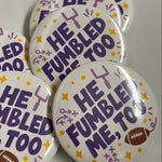 Five round pin-back buttons with a white background, featuring the text 'HE FUMBLED ME, TOO' in purple, decorated with purple and yellow stars and doodles, and one button showing a small brown football.