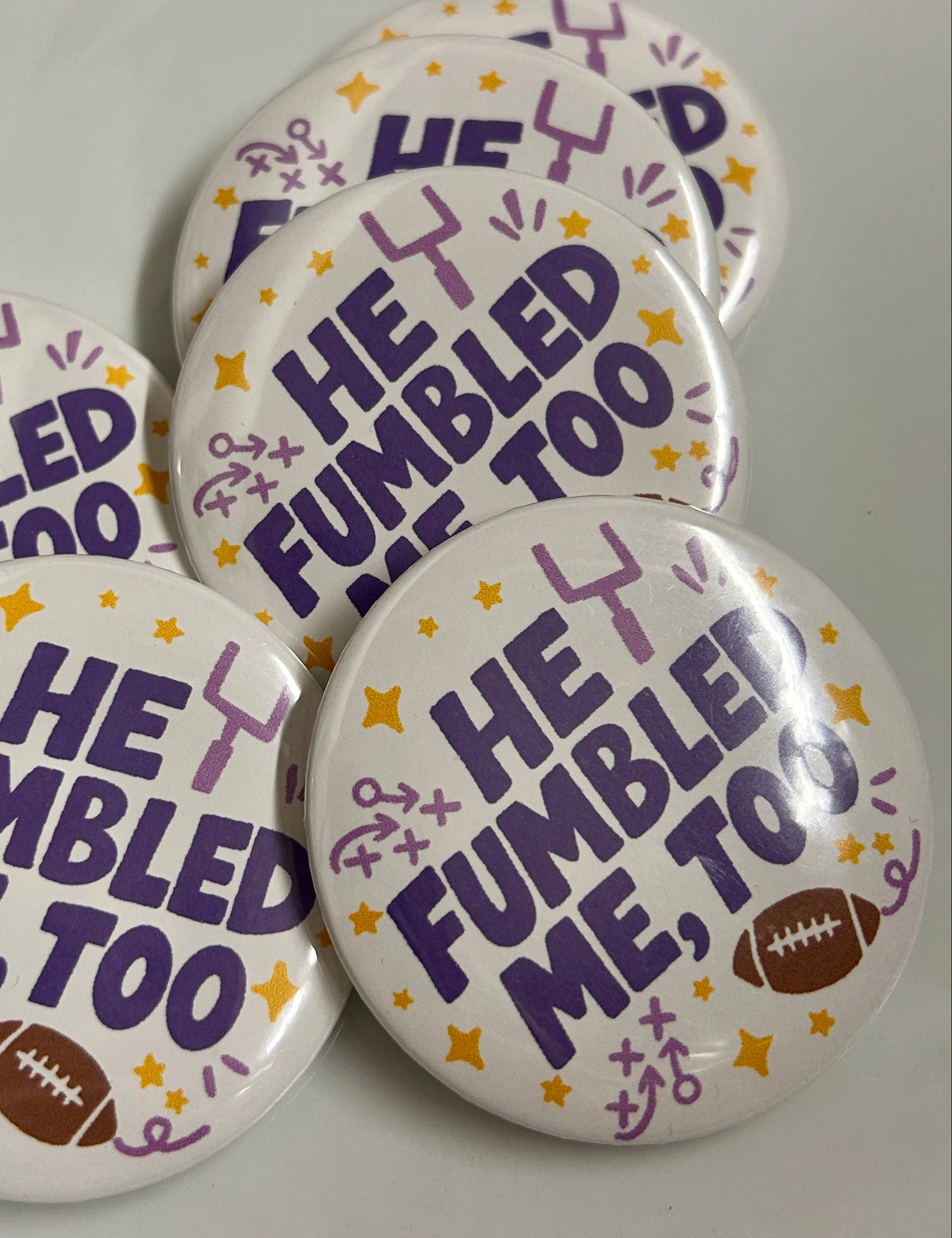 Five round pin-back buttons with a white background, featuring the text 'HE FUMBLED ME, TOO' in purple, decorated with purple and yellow stars and doodles, and one button showing a small brown football.