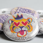 A close-up of several round pin-back buttons displayed together. The central button features a stylized tiger face with heart-shaped eyes, a purple crown, and colorful abstract details.