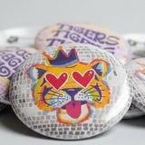 A close-up of several round pin-back buttons displayed together. The central button features a stylized tiger face with heart-shaped eyes, a purple crown, and colorful abstract details.