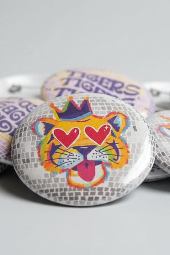 A close-up of several round pin-back buttons displayed together. The central button features a stylized tiger face with heart-shaped eyes, a purple crown, and colorful abstract details.